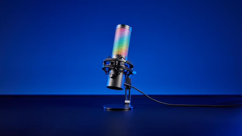 Product image of the HyperX QuadCast 2 S gaming mic. The mic (with over 100 LEDs) sits on a black desk in front of a blue background. Dramatic shadows.