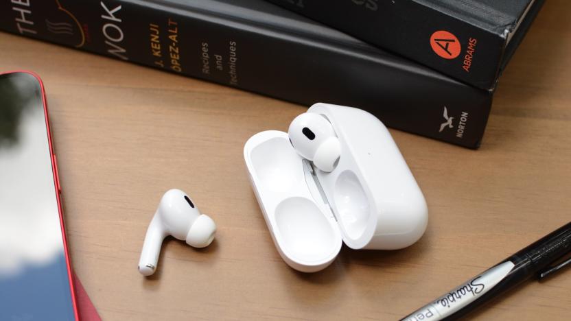 One Apple's AirPods Pro earbud sits on a desk next to an open carrying case, in which the other earbud is stored. Books, an iPhone and a pen surround the AirPods.