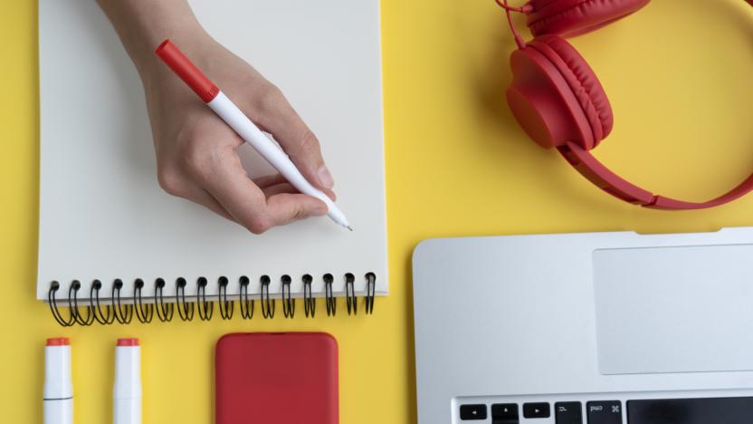 A high angle shot of a person taking notes in a notebook with headphones, pens and a laptop on a yellow surface