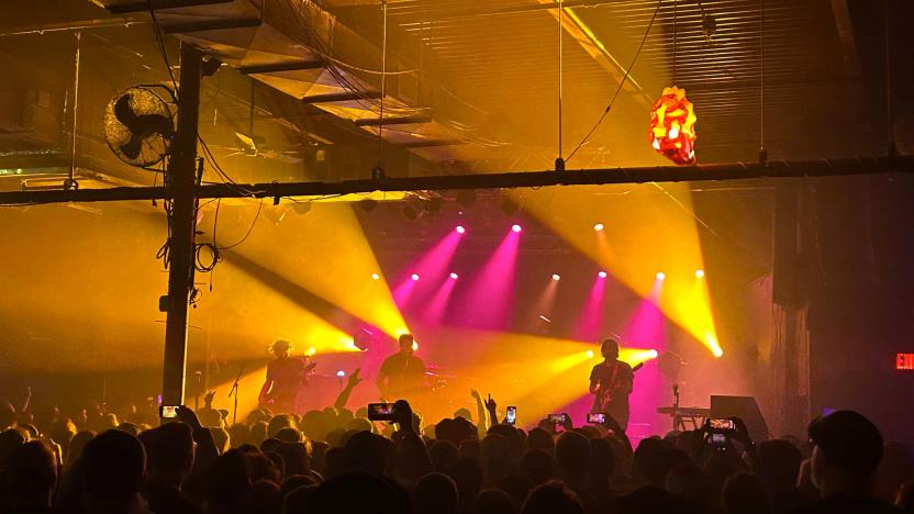 A band called Thrice is seen playing on stage from the perspective of an audience member. People are holding up phones to film them and the band is lit by pink and yellow spotlights.