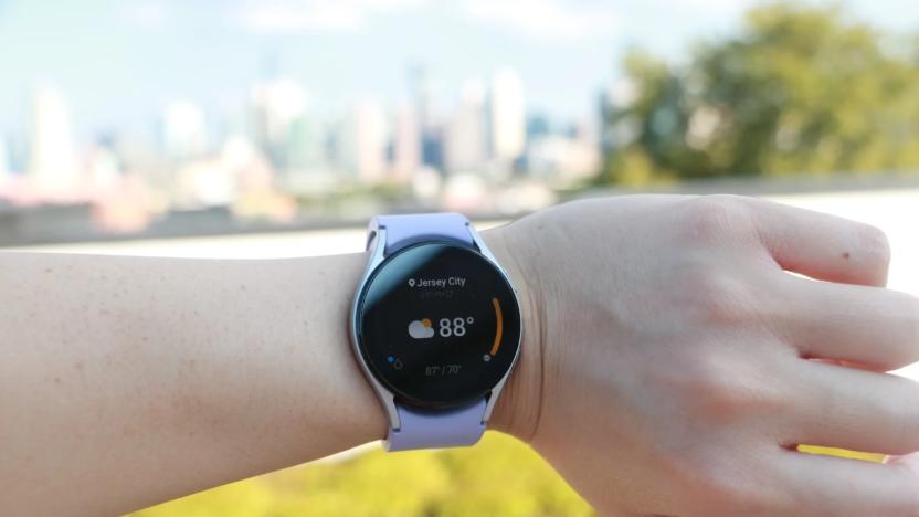 Photo of an arm wearing the Samsung Galaxy Watch 5 with lavender band against a background of trees, grass and buildings.