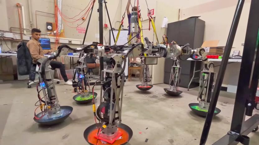 Photo inside an MIT lab of a student looking over a spider-like robot with six worm-like legs and wok-shaped “shoes.”