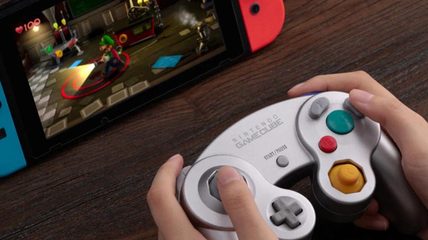 A wireless GameCube controller pictured in a person's hands, with a Nintendo Switch propped up in front of them running Luigi's Mansion