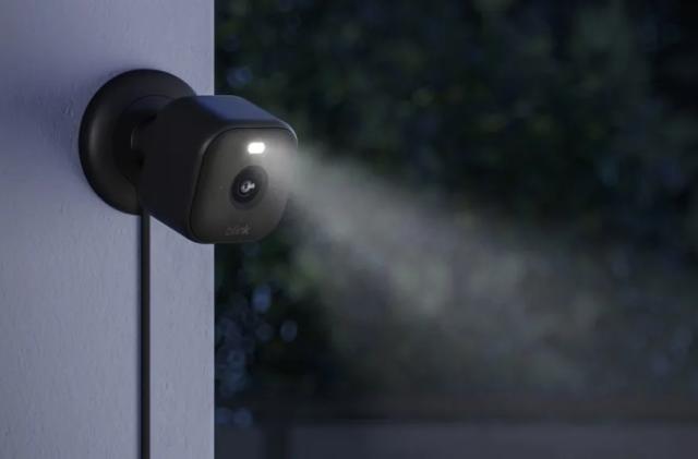 The new Blink Mini 2 camera in black shown mounted to an outside wall at nighttime with the LED spotlight shining