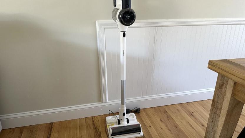 A white cordless stick vacuum sitting on its charging base. It's on wood tiled floor with light brown walls behind and the corner of a wooden table visible to the right.