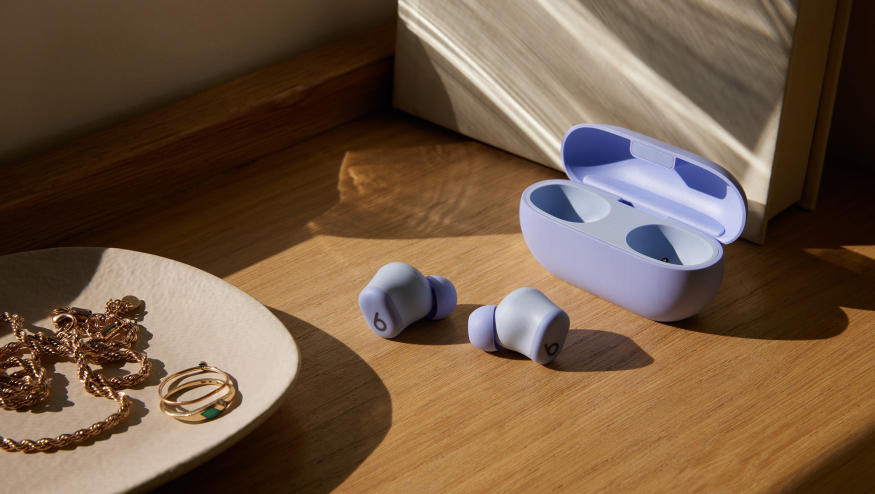 Beats Solo Buds in purple with both earbuds sitting on a wooden shelf outside of their case. A jewelry dish with rings, and other gold items sits in the foreground. 