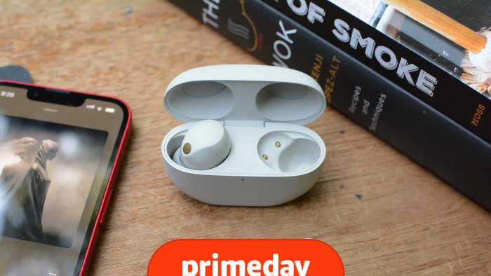 The Sony WF-1000XM5 earbuds are on a wooden table with books nearby along with a phone displaying an album cover. The prime day logo is at the bottom of the image. 