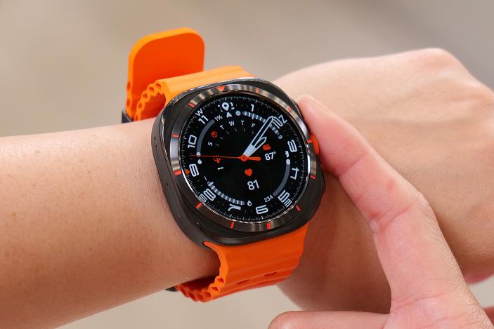 The Samsung Galaxy Watch Ultra on a wrist in mid air, showing an analog watch face with a black background and white and orange markings. A finger is resting against the middle button on the right edge.