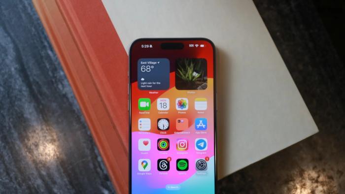 The iPhone 15 Pro Max sitting on a book on top of a desk. The phone shows its Home Screen, and the book is light gray with a reddish-orange spine. The table is a dark gray.