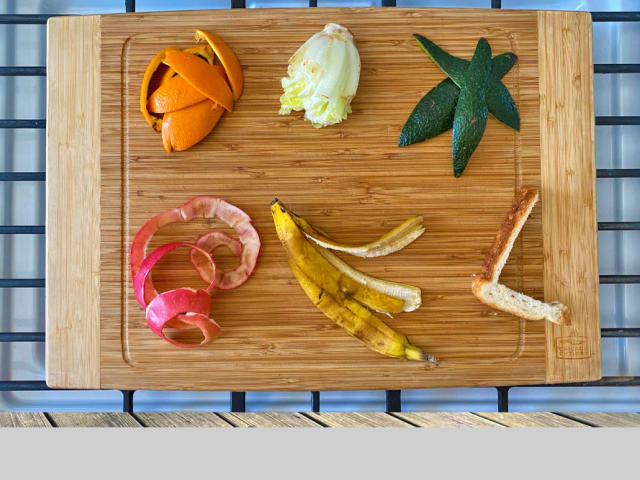 Food scraps, including lettuce, sandwich crust and peels from an orange, apple and banana, are arranged on a bamboo cutting board. 