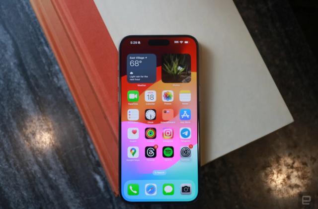 The iPhone 15 Pro Max sitting on a book on top of a desk. The phone shows its Home Screen, and the book is light gray with a reddish-orange spine. The table is a dark gray.