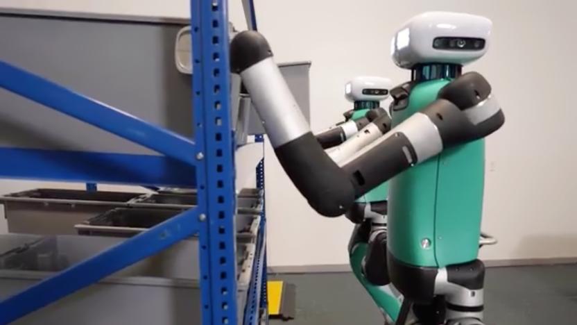 Two humanoid bipedal robots reaching onto warehouse shelves to grasp plastic storage bins.
