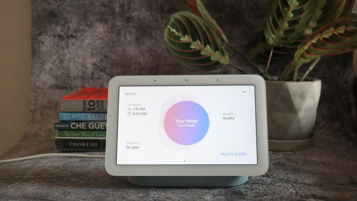 A head-on view of the Google Pixel Tablet resting on its magnetic speaker stand with a plant and small stack of books in the background.