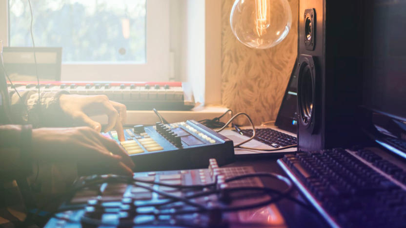 A side from of someone's home studio production setup, showing a few synths and keyboard, next to a laptop and speakers on a desktop.