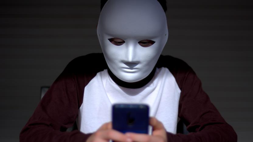 Teen boy with a white mask that makes him anonymous using the phone to make cyberbullying