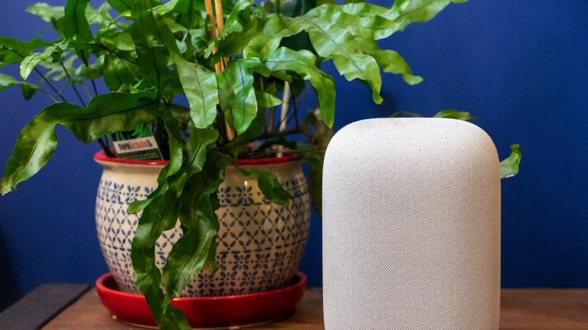 Google Nest Audio smart speaker sitting on a wooden tablet next to a green plant, against a blue wall.