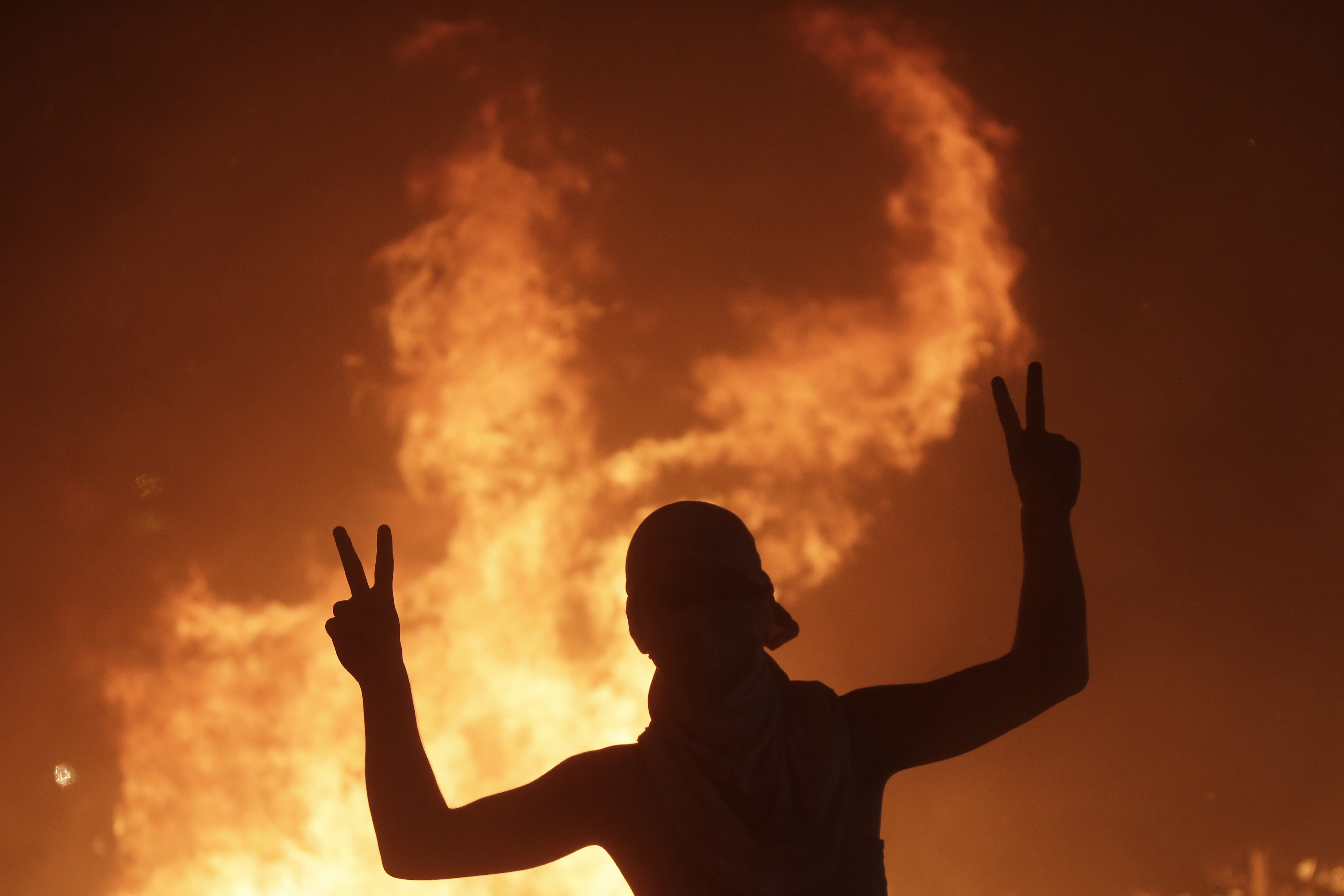 An anti-government protester makes victory signs in front a fire set to block a road during a demonstration in Beirut, Lebanon, Thursday, Oct. 17, 2019. Scores of people are protesting in Beirut and other parts of Lebanon over the government's plans to impose new taxes amid a harsh economic crisis in the country. (AP Photo/Hassan Ammar)