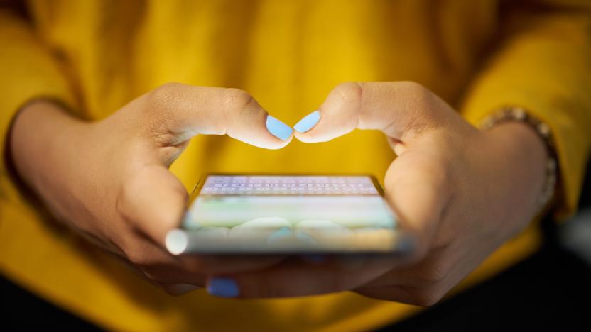 Young woman using cell phone to send text message on social network at night. Closeup of hands with computer laptop in background