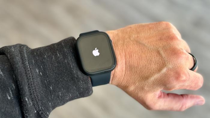 A left arm / wrist / hand wearing an Apple Watch with a white Apple logo on its otherwise black screen. Blurred gray background.