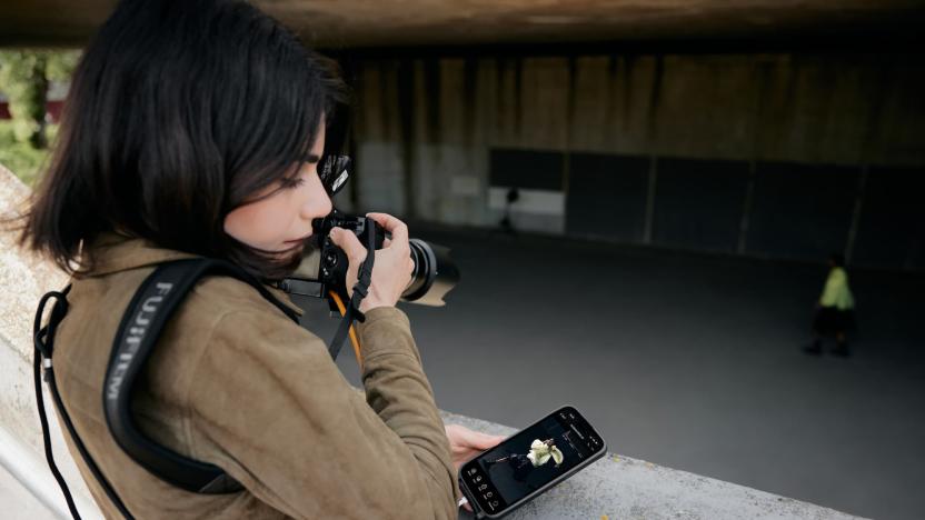 A photographer looks away from their camera toward their phone, which displays a phot-editing app.