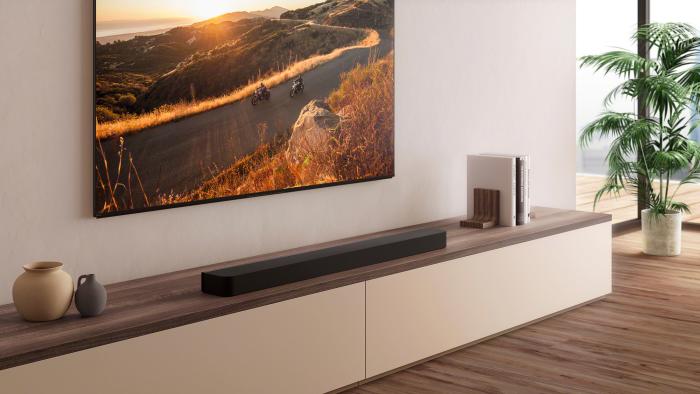 A black soundbar on a wooden shelf underneath a TV with a mountain road sunset scene on the display.
