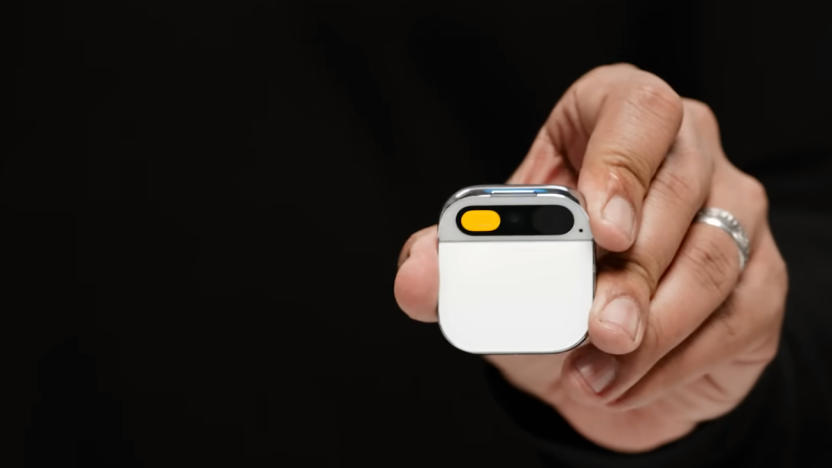 The hand of co-founder Imran Chaudhri holding the Humane AI Pin. Closeup. The small device has a white face, and is square with round corners and a section at the top for a camera.