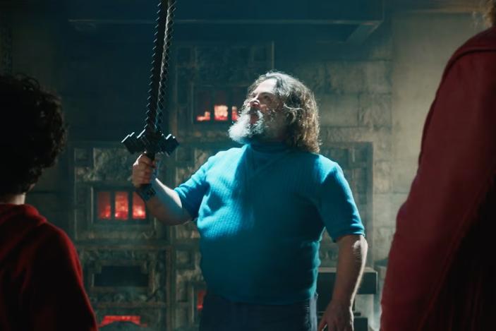 A still from A Minecraft Movie showing Jack Black wearing a blue shirt as the iconic character Steve, holding a crafted sword. We see two other characters from the back, both wearing red and looking at Jack Black