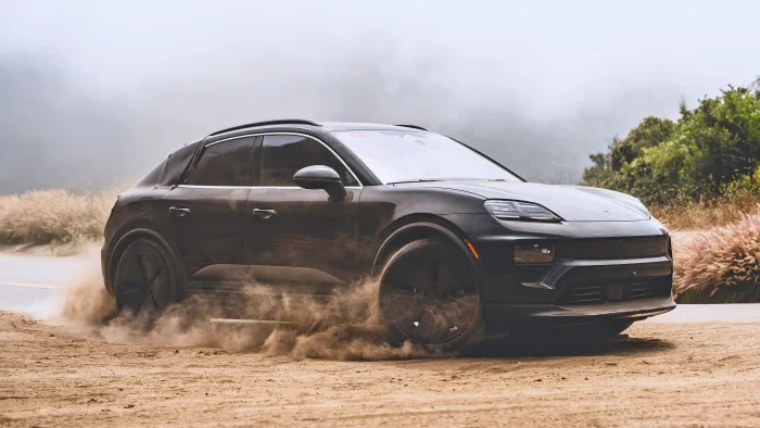 A black Porsche Macan 2025 EV speeds through the dirt shoulder of a road kicking up small dust clouds.