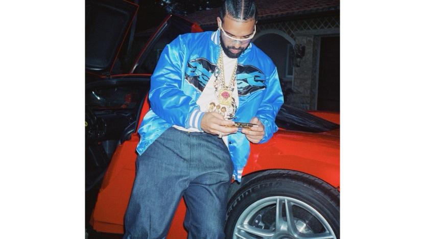 The rapper Drake leaning against a red car, looking at his phone.