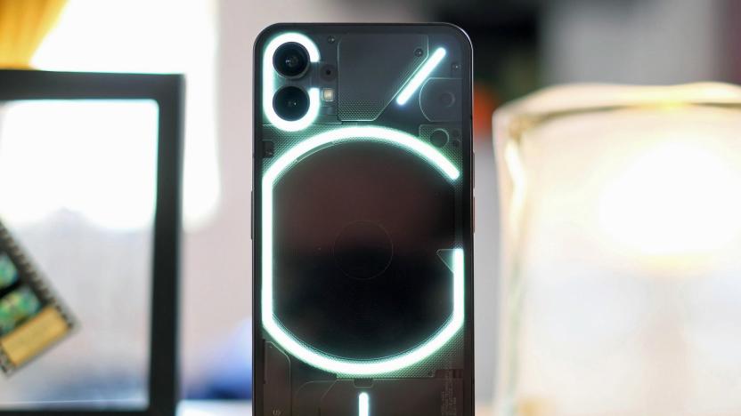 The new Nothing Phone 1 stands on its end on a table. The phone is black with a see-through back showing a glowing series of lines and rings.
