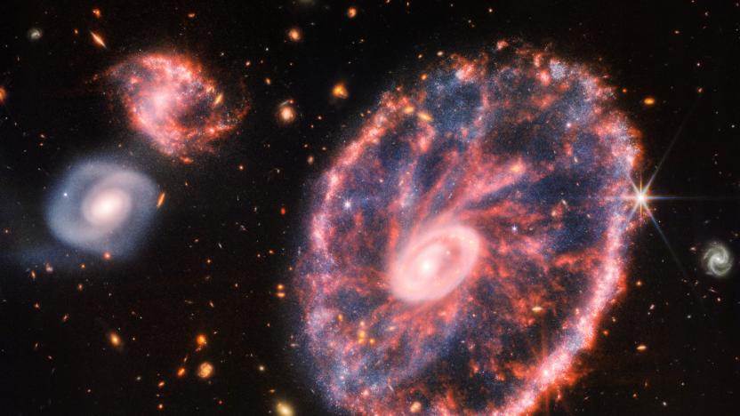 A large pink, speckled galaxy resembling a wheel with with a small, inner oval, with dusty blue in between on the right, with two smaller spiral galaxies about the same size to the left against a black background.