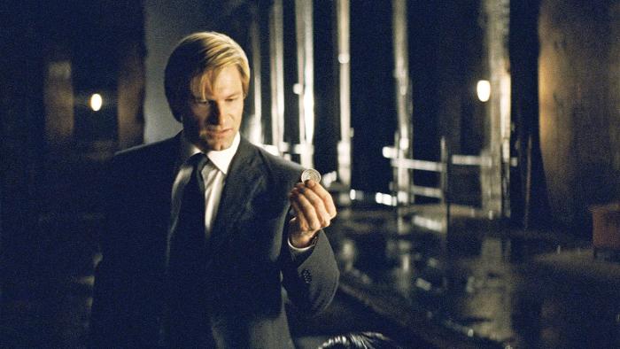 Promotional still from the movie "The Dark Knight" showing Harvey Dent (Aaron Eckhart, pre-Two-Face transformation) holding a coin before he flips it. He’s in a dark warehouse.