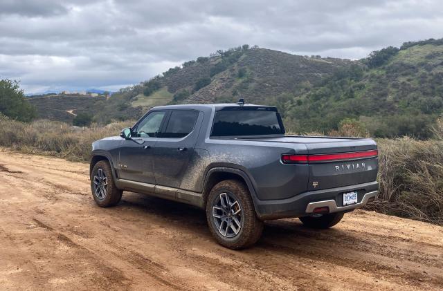 Photo from behind and to the left of the Rivian R1T electric pickup truck. It drives on a dirt road in green hilly terrain.