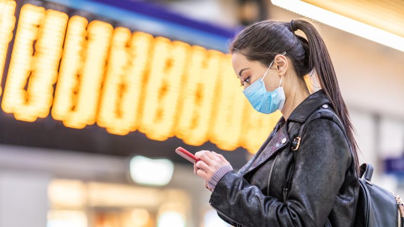 Chinese woman wearing face mask at train station to protect from smog and virus - young asian woman looking at her smartphone with departure arrivals board behind - health and travel concepts