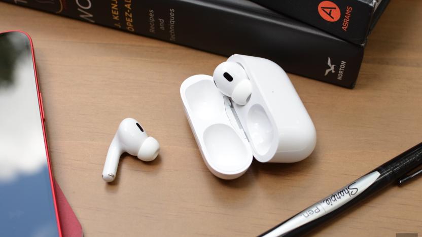 One Apple's AirPods Pro earbud sits on a desk next to an open carrying case, in which the other earbud is stored. Books, an iPhone and a pen surround the AirPods.