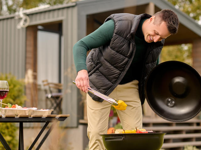 Smiling man leaning over the open brazier and gripping the slice of bell pepper with barbecue tongs