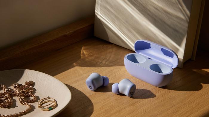 Beats Solo Buds in purple with both earbuds sitting on a wooden shelf outside of their case. A jewelry dish with rings, and other gold items sits in the foreground. 