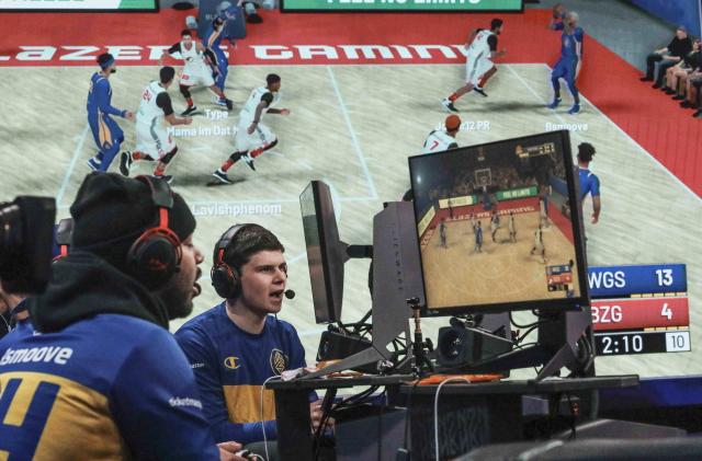 A giant monitor shows play as Warriors Gaming Squad teammates Charles "CB13" Bostwick, center, from New York, and his teammate Alexander Reese, left, from Milwaukie, Or., react to scoring during the NBA 2k League (NBA2KL) professional esports playoffs, Wednesday, July 24, 2019, in Queens borough of New York. The teams kicked off day one of the NBA2KL playoffs with Blazers on the losing 67-45. (AP Photo/Bebeto Matthews)