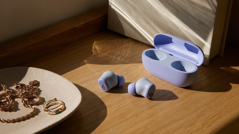 Beats Solo Buds in purple with both earbuds sitting on a wooden shelf outside of their case. A jewelry dish with rings, and other gold items sits in the foreground. 