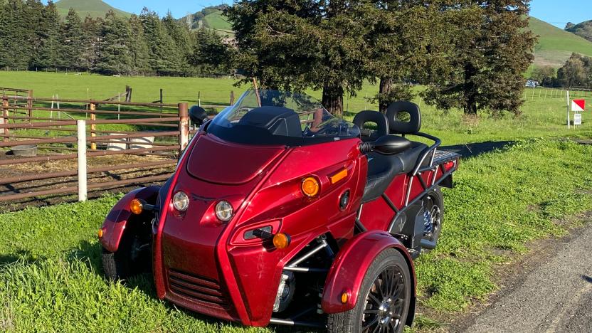 The Arcimoto three-wheel EV parked on the side of a dirt road next to a farm.