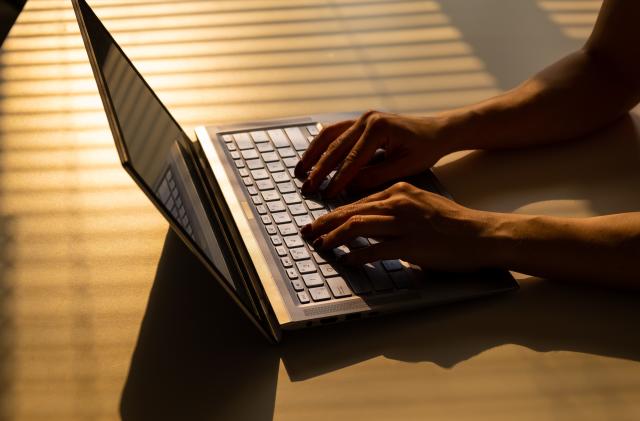 A woman is typing on a laptop keyboard on a white table. The shadow from the blinds falls on the desktop