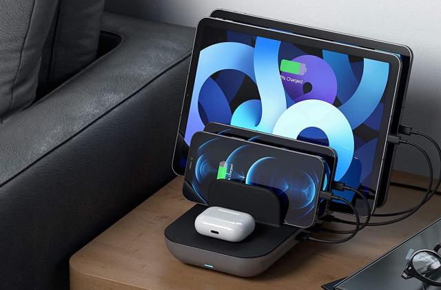 A photo of Satechi's Dock5 Multi-Device Charging station, depicting the multi-port charger as it powers up several iPhones, iPads, and AirPods on top of a brown side table, next to the arm of a black leather couch. 