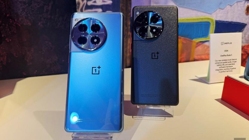 Two OnePlus 12 phones site on display stands at an event. Both have their rear camera facing forward, one is a light blue metallic and the other is a dark grey.