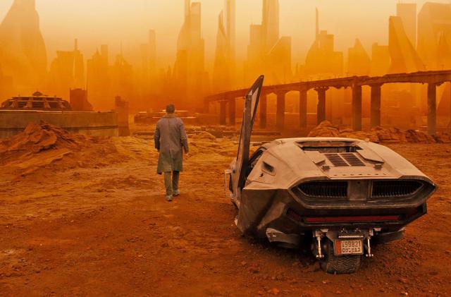 A promo still image from Blade Runner 2049 with the main character walking away from his vehicle into  an abandoned derelict city that's overcast with an orange glow.