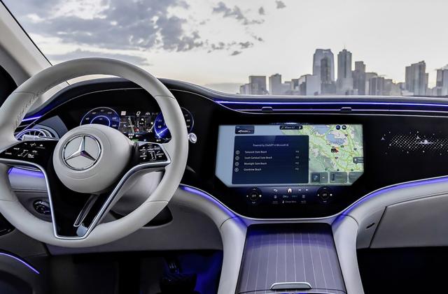 An in-car view of a modern Mercedes interior. The steering wheel is on the left, with main console in the center. In the background is a city skyline.