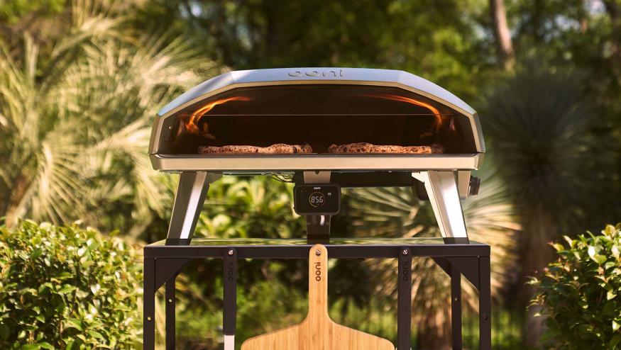 Ooni Koda 2 Max pizza oven sitting on a metal stand in an outdoor setting. Two pizza peels are hanging on the front of the stand and there's visible fire inside the pizza oven.