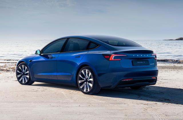 A blue Tesla Model 3 (2024) is parked on a sandy beach right near the edge of where the tide is coming in.