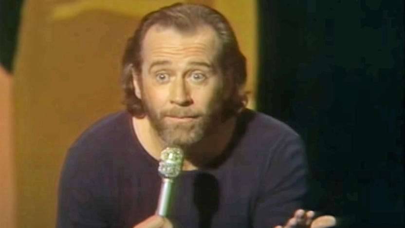 George Carlin holds a microphone and eyeballs the crowd in a questioning manner in the middle of doing a bit on stage in the 1970s.