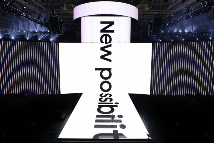 Stage with huge ultrawide screen on the back, and a perpendicular screen stretching above and below it onto the runway. "New possibilities" on the screen.