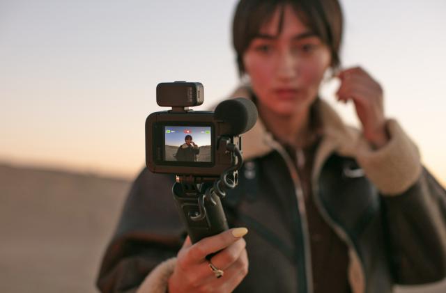 A woman takes a selfie using the GoPro Volta Battery Grip while standing on a beach or with a sand dune behind her.